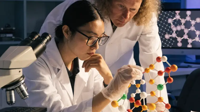 Two scientists in white lab coats examining a complex, multi-colored molecular model in a research laboratory setting.