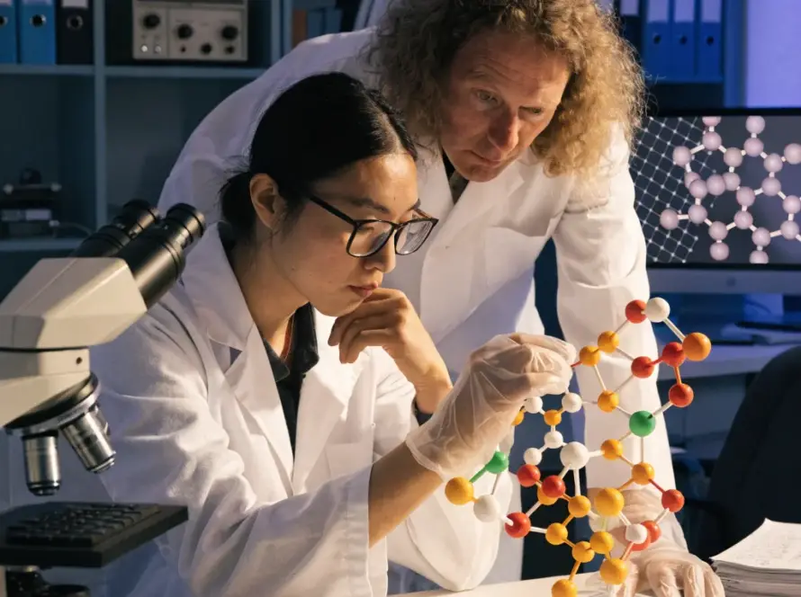 Two scientists in white lab coats examining a complex, multi-colored molecular model in a research laboratory setting.